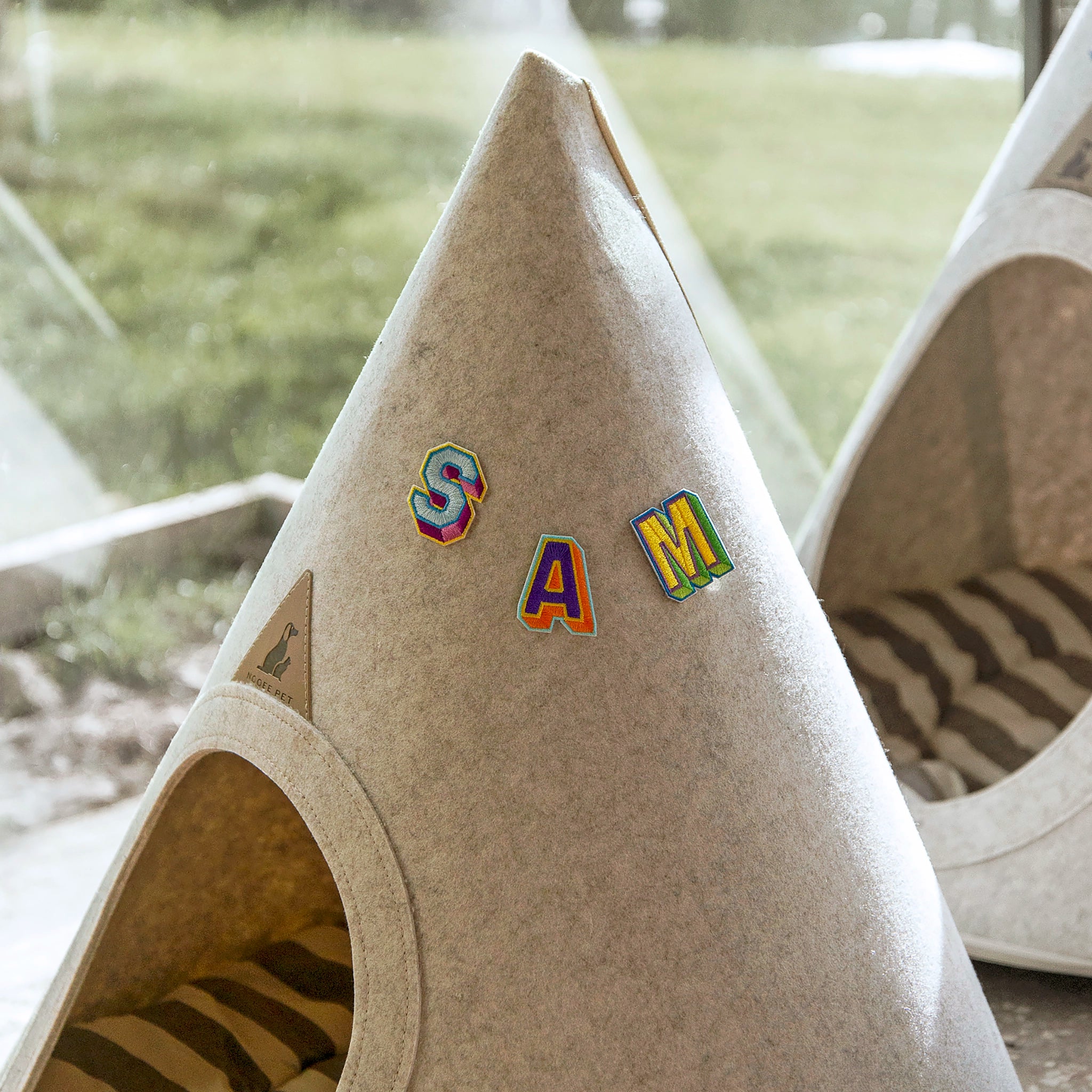 Nooee Pet teepee with colorful embroidered alphabet patches spelling “SAM” on the front, placed indoors near a window with a view of green grass outside.