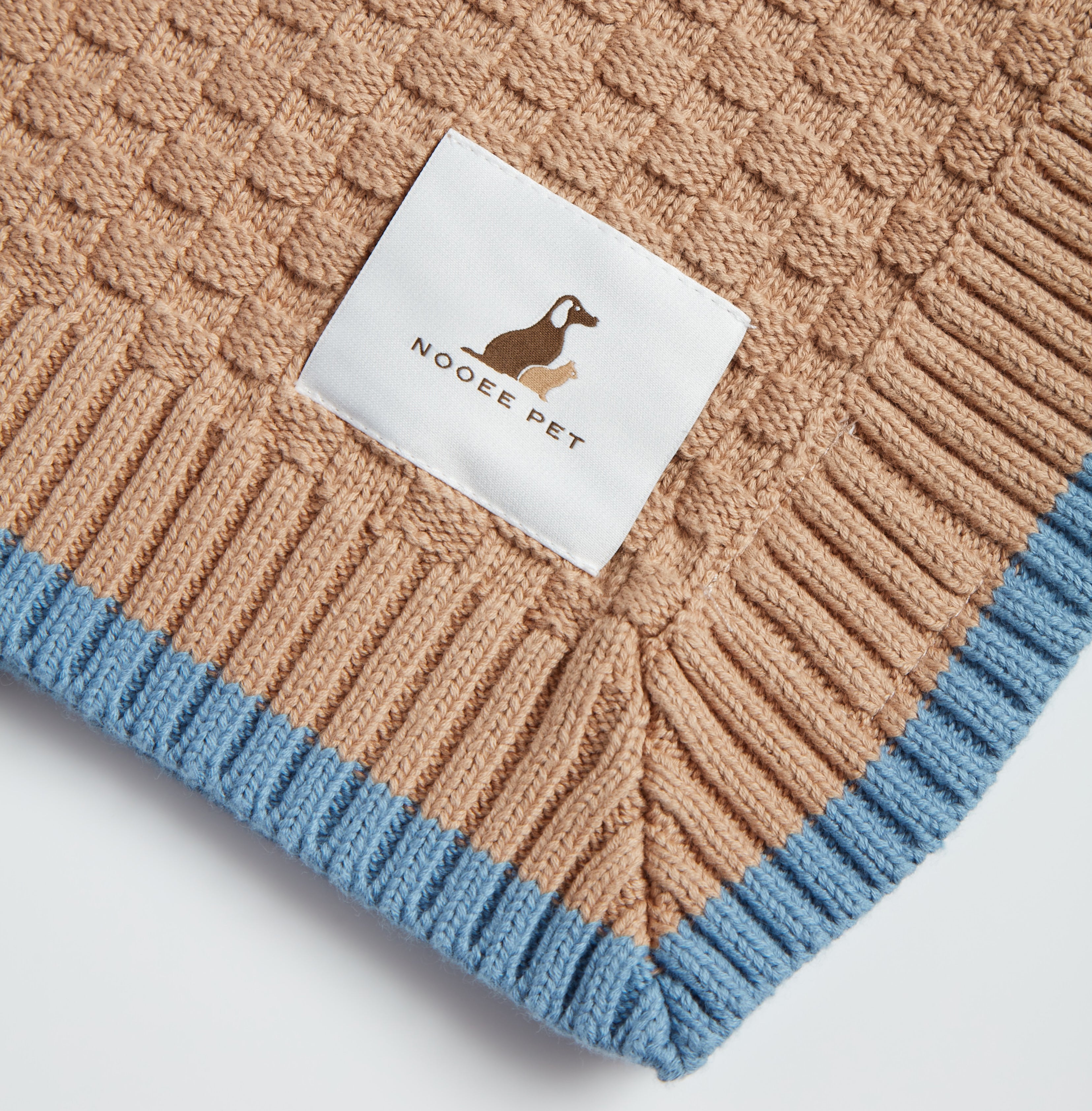 Close-up of Nooee Pet logo tag on the corner of a brown organic cotton blanket with blue ribbed trim and a waffle-knit texture.