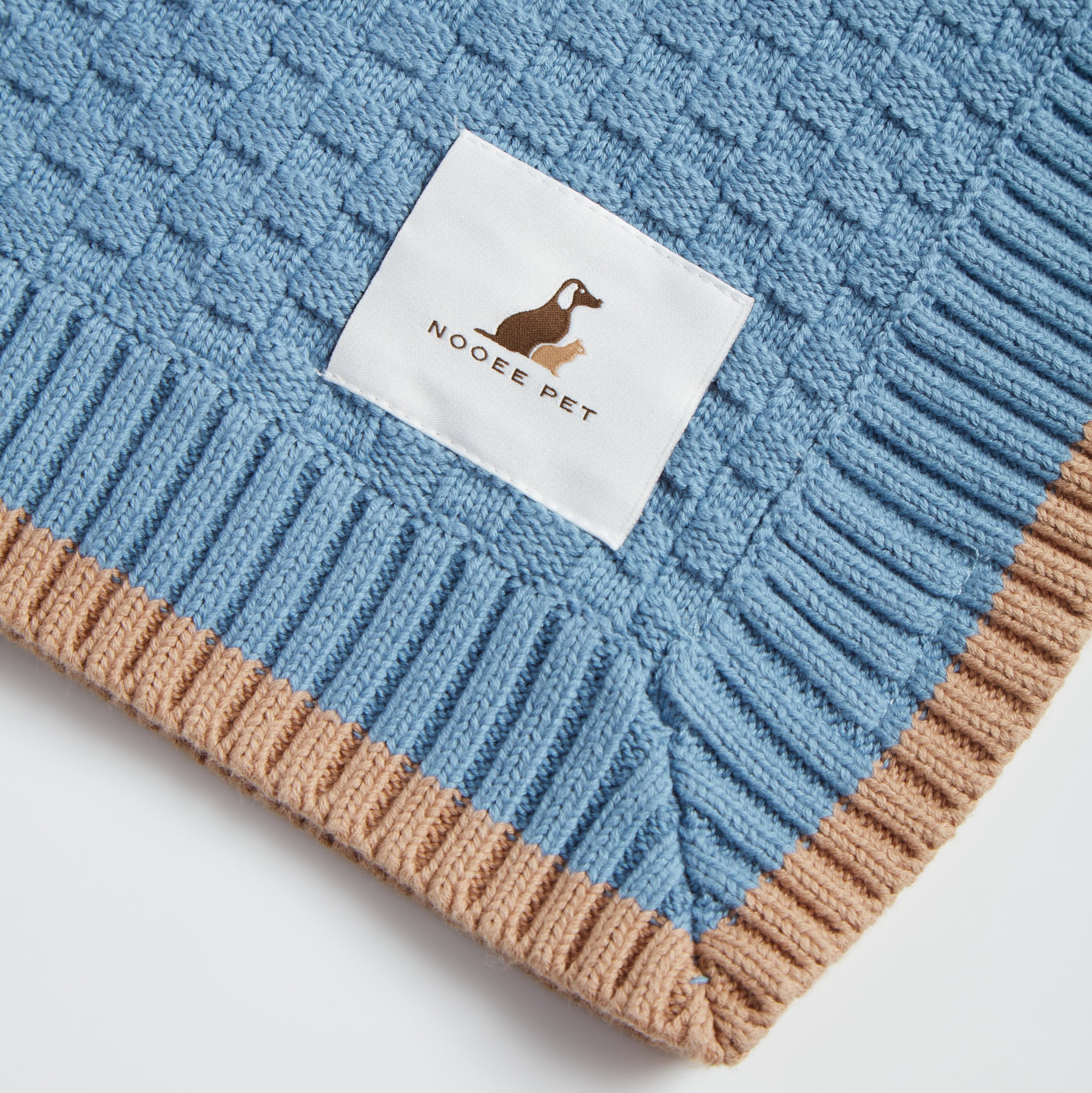Close-up of Nooee Pet logo tag on a blue organic cotton blanket with beige ribbed trim and a waffle-knit texture.
