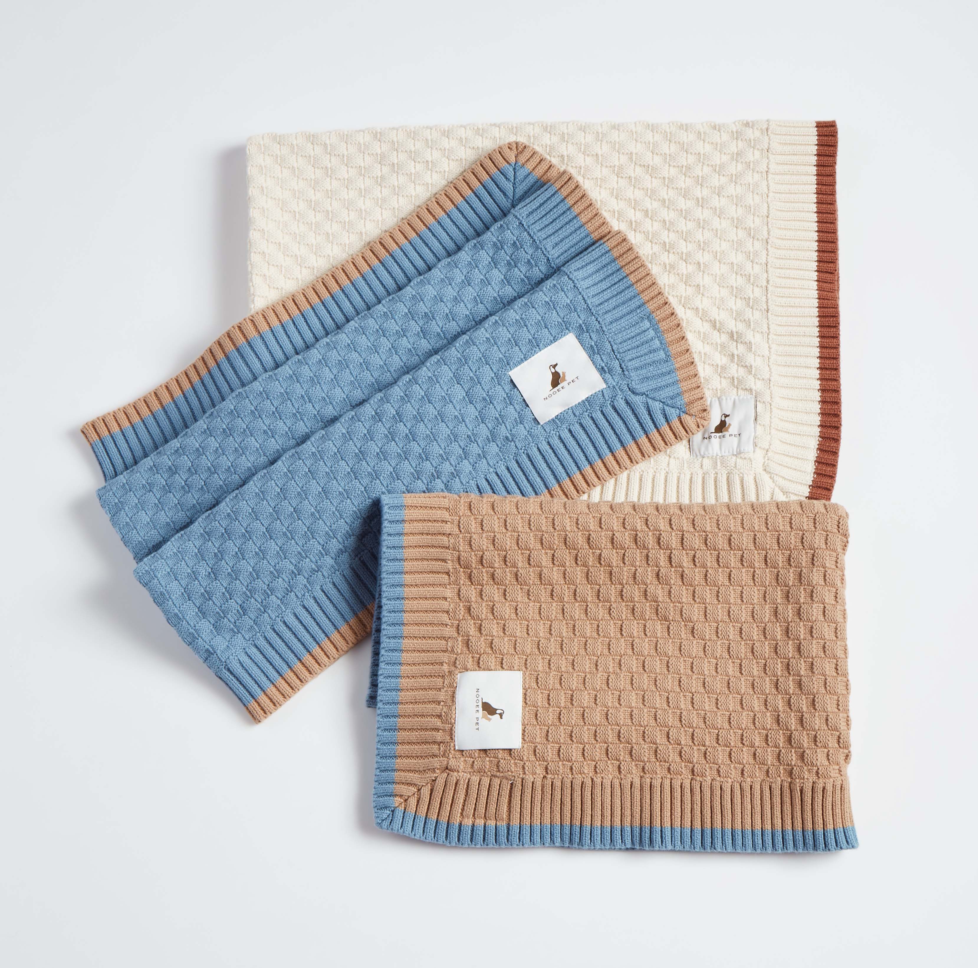 Three neatly folded Nooee Pet Organic Cotton Blankets in cream, blue, and tan colors with contrast ribbed trim, showcasing the soft texture and woven pattern.