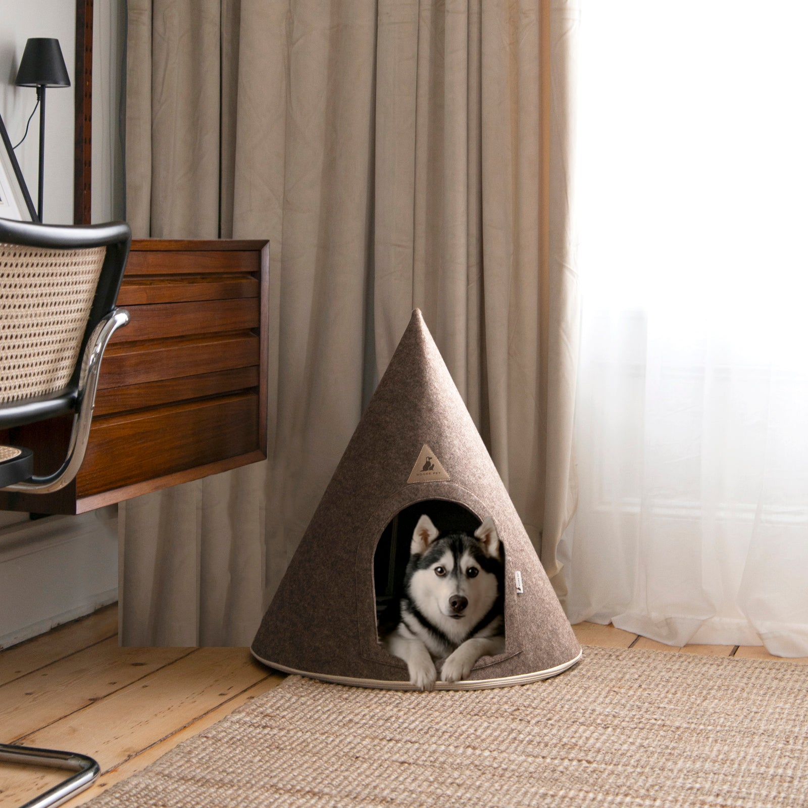 Husky sitting inside a brown felt pet cave in a cozy room with wood flooring, curtains, and a mid-century desk chair.