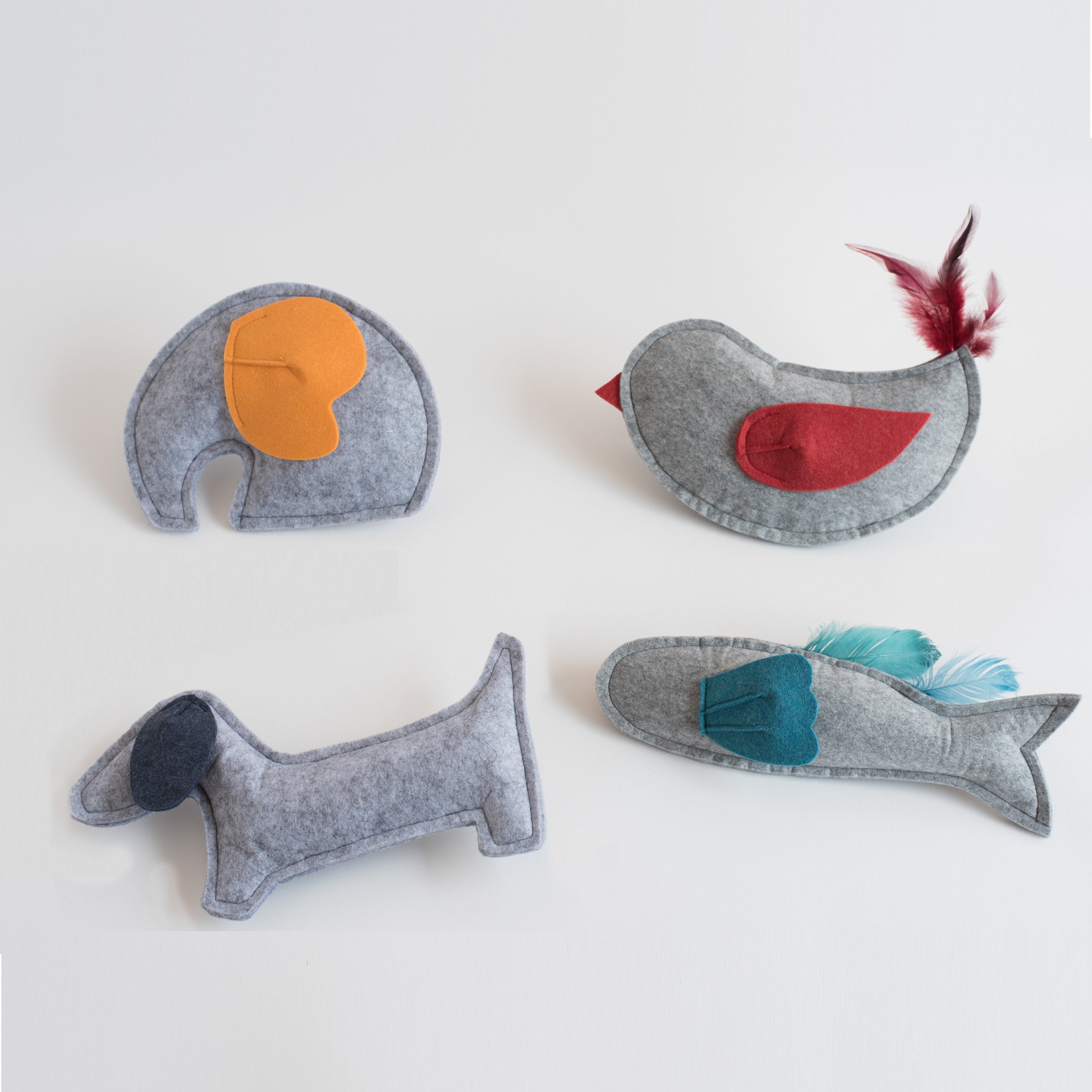 Four felt animal-shaped pet toys in various colors and designs, including a gray dog, elephant, fish with feathers, and bird with red wing and feathers, arranged on a white background.