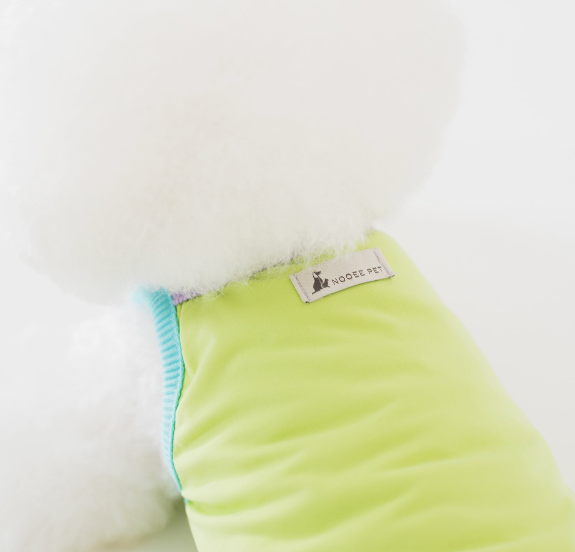 Close-up of a white fluffy dog wearing the Nooee Pet CoolPaws Summer Shirt in light green with aqua blue trim and a visible Nooee Pet logo tag on the back.