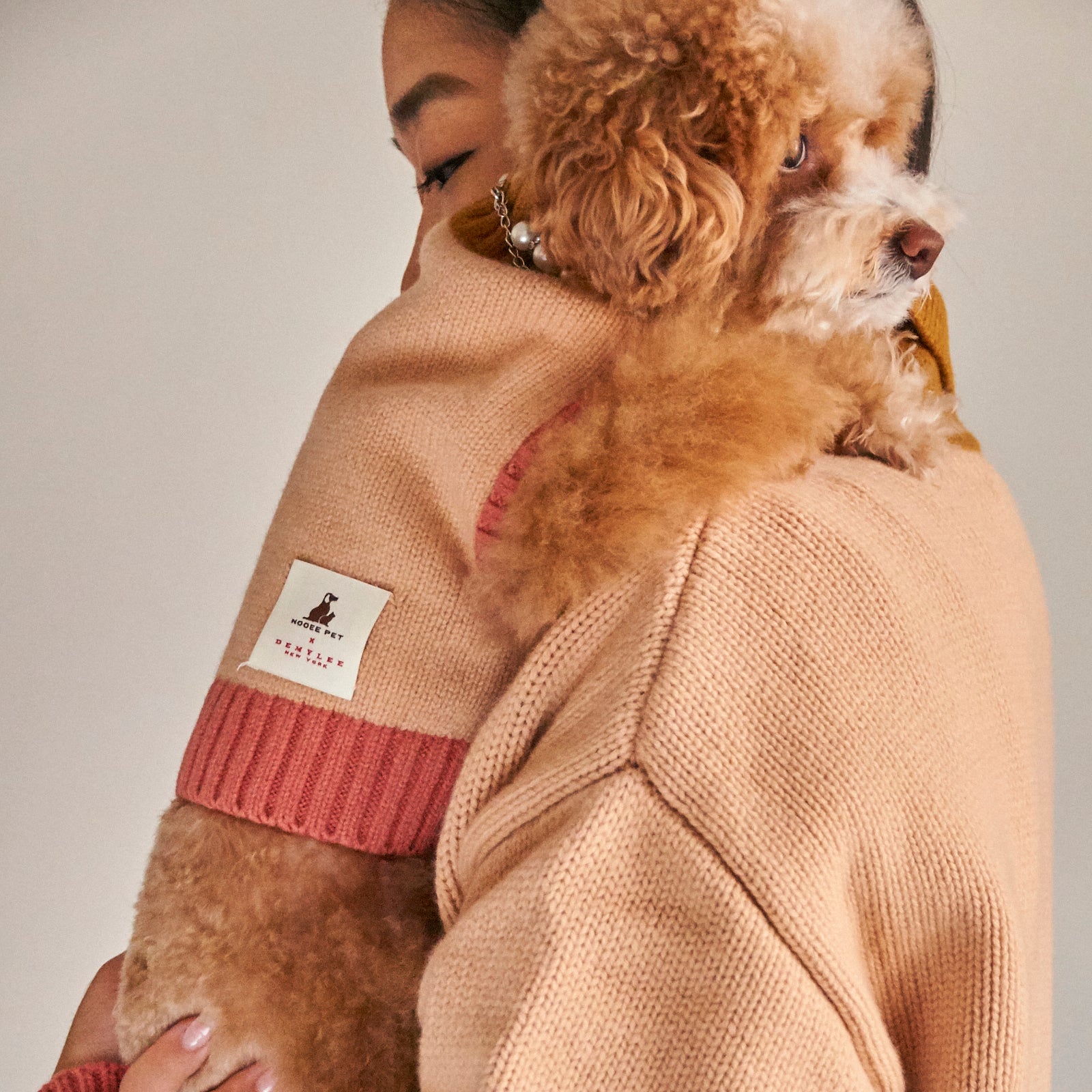 A small poodle wearing the Nooee Pet x DEMYLEE fawn merino wool dog sweater with pink ribbed trim, being lovingly held by a person in a matching knit outfit.