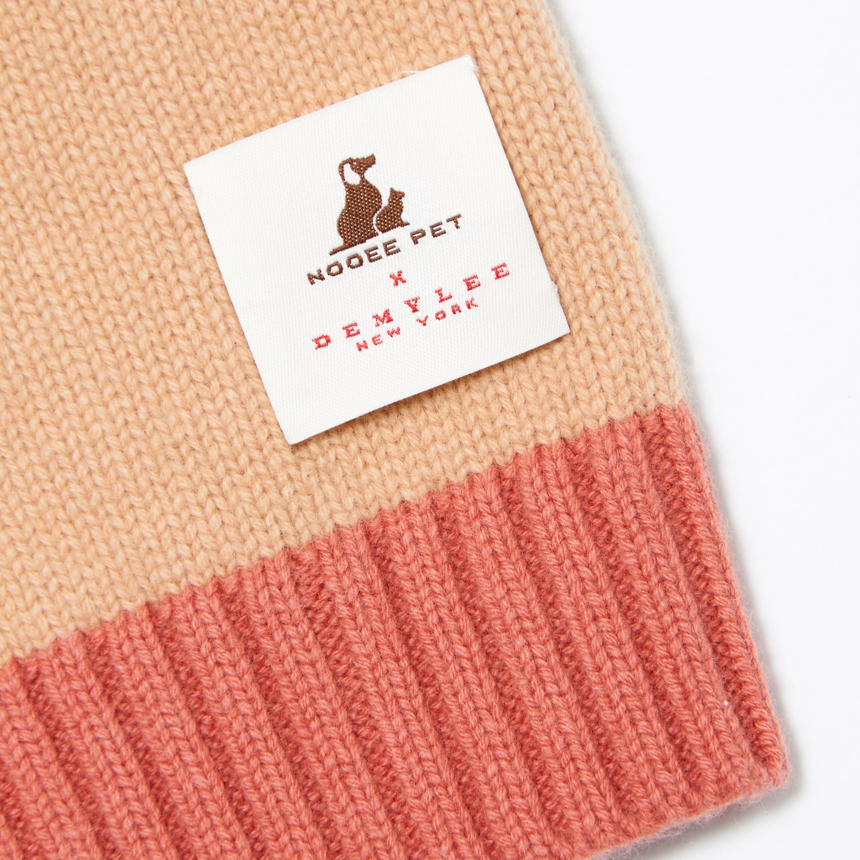 Close-up of the Nooee Pet x DEMYLEE New York label stitched onto the hem of the fawn sweater, showcasing the pink ribbed knit detail and beige body of the garment.