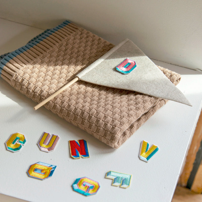 Colorful embroidered alphabet patches displayed on a white surface, with a gray felt flag showcasing the letter "D" and a folded taupe knit blanket underneath.