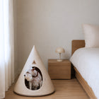 A small white dog sits inside the Nooee Pet dog cave with an oval opening, placed on a wooden floor beside a bed and bedside table in a softly lit bedroom.
