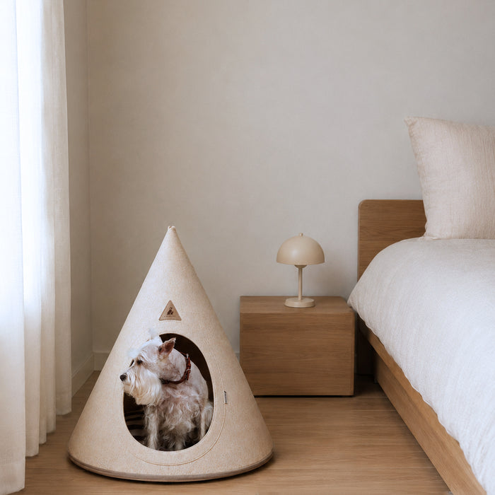 A small white dog sits inside the Nooee Pet dog cave with an oval opening, placed on a wooden floor beside a bed and bedside table in a softly lit bedroom.