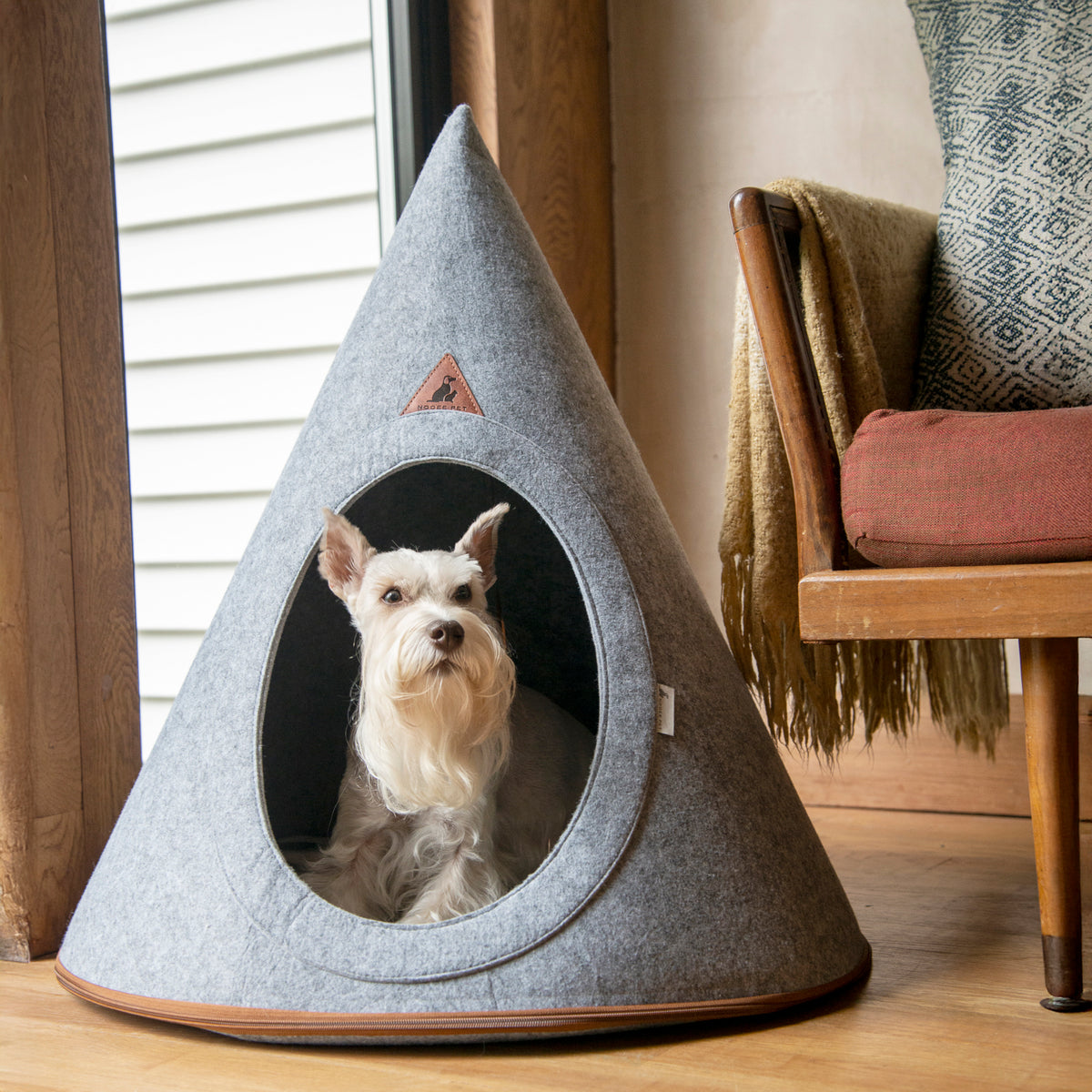 Gray felt pet cave with a small white dog sitting inside, styled beside a wooden chair in a warm home interior.