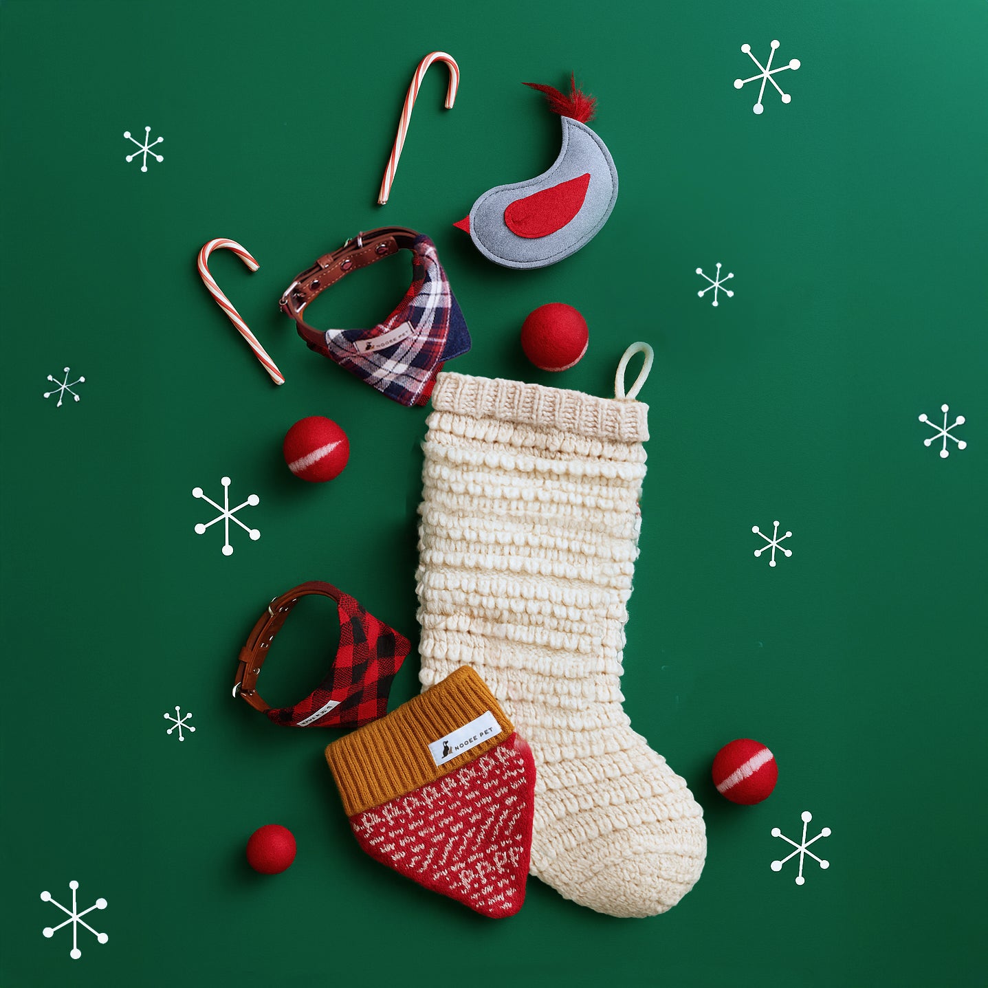 Holiday stocking stuffer gift layout featuring Nooee Pet wool toy balls, felt animal toy, neck warmers, and patterned collars arranged on a green backdrop with a cream knitted stocking.