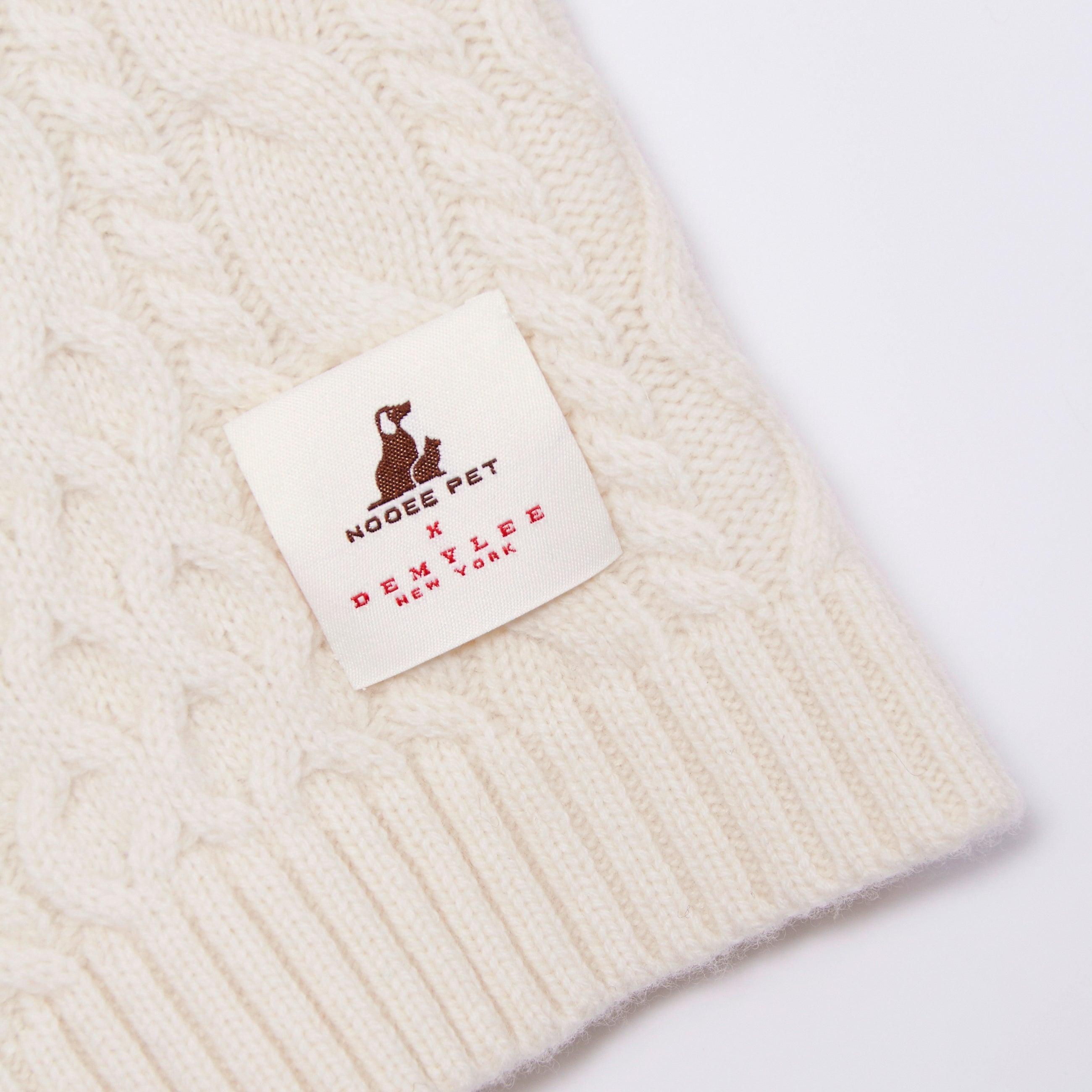 Close-up of the Nooee Pet x DEMYLEE New York collaboration label stitched onto the off white wool cable dog sweater.